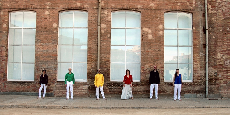 Six members of Forum Neue Vokalmusik in white trousers and coloured shirts, standing against a brick wall with large windows