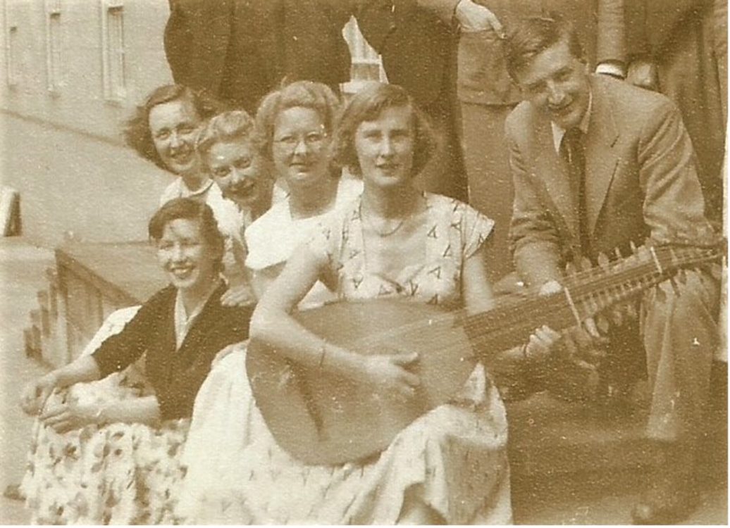 A sepia photo from the 1950s of people sitting on steps. One is holding a lute